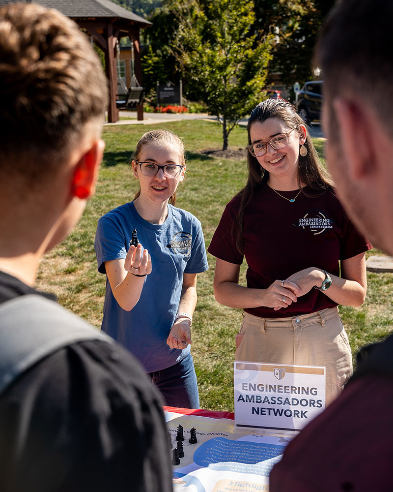 Two students represent Geneva College’s Engineering Ambassadors Network at an outdoor booth, interacting with peers and displaying materials including a chess piece and an info sign.